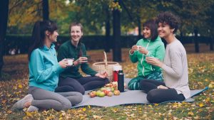 Happy girls are having picnic in park sitting on yoga mats and eating after outdoor practice in autumn, girls are talking and laughing. Communication, people and food concept.